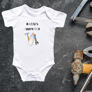 Op de afbeelding: Witte babybody met de tekst "Daddy's Apprentice" en illustraties van een hamer, sleutel en babyfles. De body is omringd door diverse gereedschappen, waaronder een zaag, tang en verstelbare moersleutel, op een donkergrijze ondergrond.