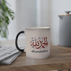 Könnte beinhalten: Eine weiße Keramiktasse mit schwarzem Henkel, mit burgunderfarbener arabischer Kalligraphie und der englischen Übersetzung "Alhamdulillah" in Schwarz. Die Tasse steht auf einer Holzoberfläche, mit einer Zeitung und einer grauen Teekanne im Hintergrund.