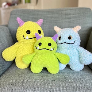 Könnte beinhalten: Drei gehäkelte Plüschtiere in Form von Monstern. Das größte Monster ist gelb mit lila Hörnern und einem zahnigen Grinsen. Das mittlere Monster ist grün mit lila Hörnern und einem zahnigen Grinsen. Das kleinste Monster ist hellblau mit weißen Hörnern und einem zahnigen Grinsen.
