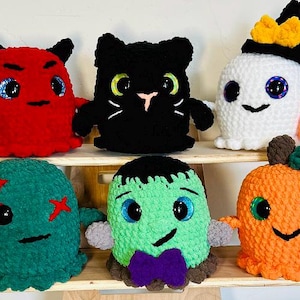 May include: A collection of seven crocheted Halloween-themed plush toys. The toys include a red devil, a black cat, a white ghost, a green monster, an orange pumpkin, and two smaller green monsters. The toys are all handmade and have big eyes and friendly faces.