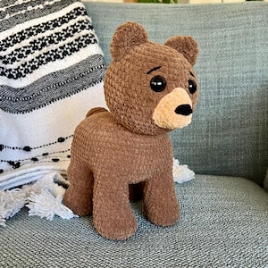 Puede incluir: Un oso de peluche de crochet marrón con ojos negros y una nariz marrón. El oso está de pie en un sofá azul claro.