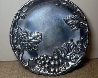 Puede incluir: Una bandeja redonda de metal plateado con un diseño de uvas y hojas. El borde de la bandeja está adornado con un motivo de vid. El centro de la bandeja presenta un racimo de uvas y hojas detalladas.