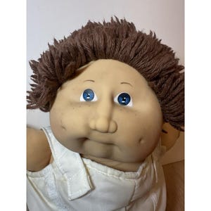 May include: A vintage doll with a soft, tan face, blue eyes, and freckles. The doll has brown yarn hair and is wearing a white outfit. The doll's facial expression is slightly puckered.