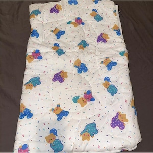 Könnte beinhalten: Weiße gesteppte Decke mit einem bunten Muster aus Teddybären, die Pyjamas tragen und Ballons halten. Der Hintergrund ist weiß mit Konfetti.