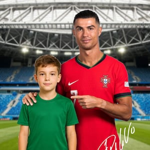 May include: A man wearing a red soccer jersey with the Portuguese flag on the chest, is standing with his arm around a young boy wearing a green soccer jersey. The man is pointing to the number 7 on his jersey. The man's signature is visible on the jersey. The background is a blurry image of a soccer stadium.