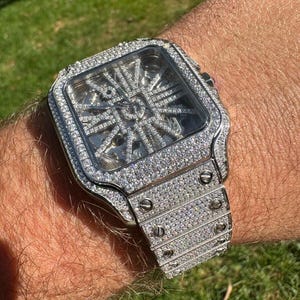 May include: A silver watch with a square face and a diamond-encrusted band. The watch face is clear and shows the inner workings of the watch.
