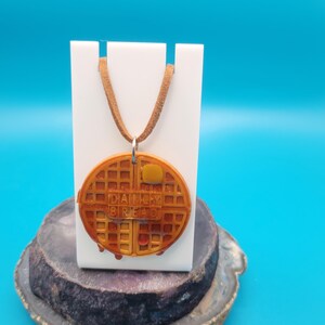 May include: A brown leather cord necklace with a waffle-shaped pendant. The pendant is brown and yellow and has the words "Daily Bread" printed on it.