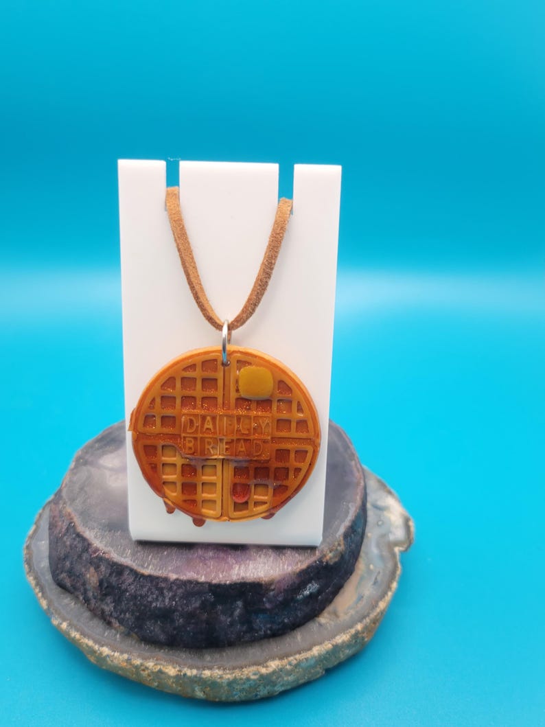 May include: A brown leather cord necklace with a waffle-shaped pendant. The pendant is brown and yellow and has the words "Daily Bread" printed on it.
