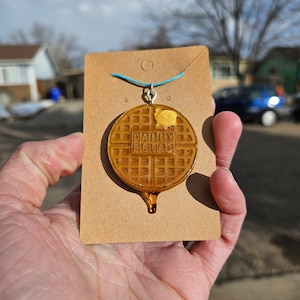 May include: A brown waffle necklace with the words "Daily Breakfast" printed on it. The necklace is on a brown card with a blue string attached to it.