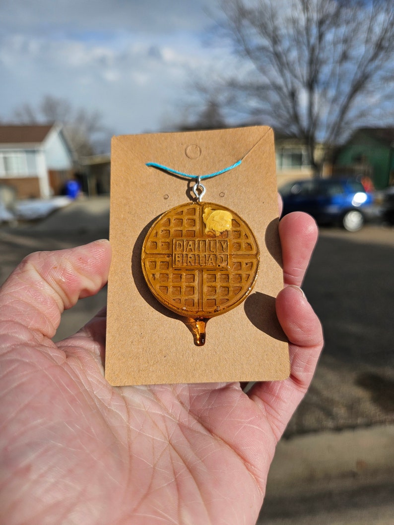 May include: A brown waffle necklace with the words "Daily Breakfast" printed on it. The necklace is on a brown card with a blue string attached to it.