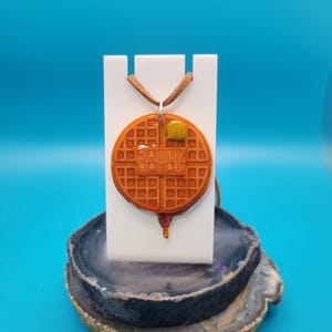 May include: A brown waffle necklace with a yellow butter pat on top. The waffle has the words "Daily Bread" written on it. The necklace is on a white stand and a gray and brown agate base.