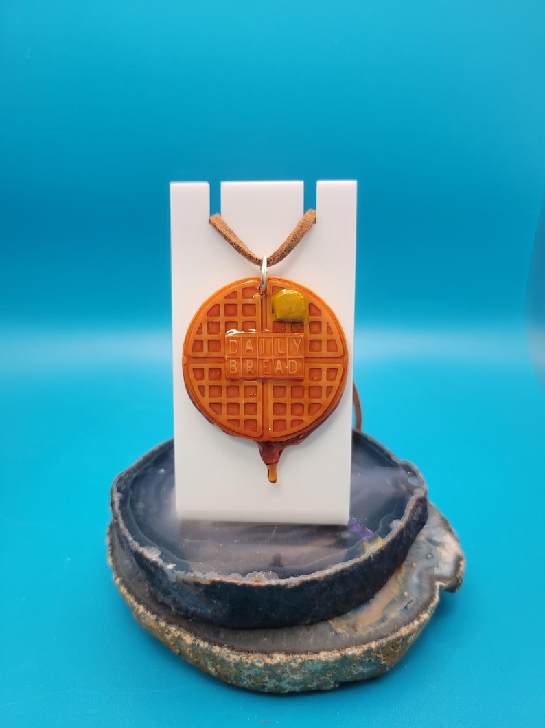 May include: A brown waffle necklace with a yellow butter pat on top. The waffle has the words "Daily Bread" written on it. The necklace is on a white stand and a gray and brown agate base.