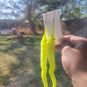 May include: A pair of neon yellow worm earrings with silver hooks. The earrings are made of fuzzy material and have a slight bend in the middle.