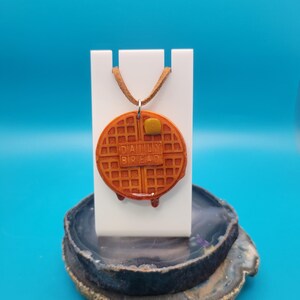 May include: A brown waffle necklace with the words "Daily Bread" on it. The necklace is on a white stand and a brown agate slice.
