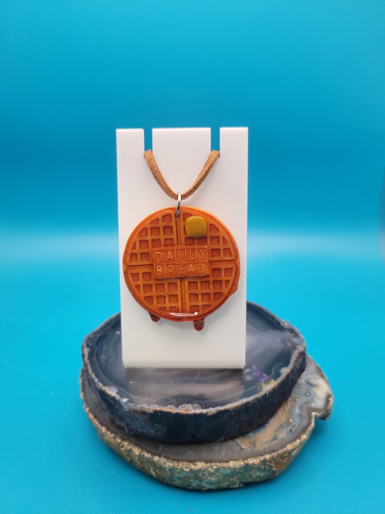 May include: A brown waffle necklace with the words "Daily Bread" on it. The necklace is on a white stand and a brown agate slice.