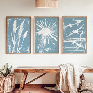Puede incluir: Tres impresiones artísticas enmarcadas con fondo azul claro y diseños blancos. Las impresiones presentan un diseño botánico, un sol radiante y aves en vuelo. Los marcos son de madera clara, sobre un banco de madera.