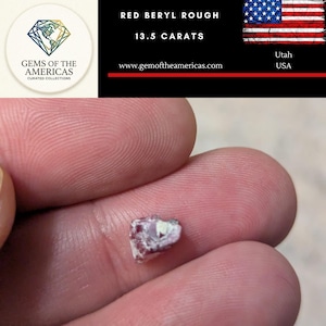 May include: A small, rough-cut red beryl gemstone, weighing 13.5 carats, is held between fingers. The image includes a logo for "Gems of the Americas" and text indicating the gem's origin in Utah, USA.
