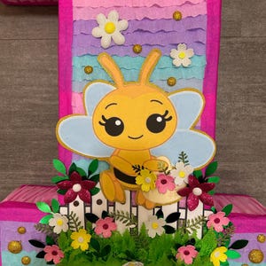 Pu&ograve; includere: Una pi&ntilde;ata rosa e blu a forma di numero uno con un'ape di cartone animato che tiene un cestino di fiori. L'ape &egrave; gialla con strisce nere e ha occhi grandi. La pi&ntilde;ata &egrave; decorata con fiori e glitter.