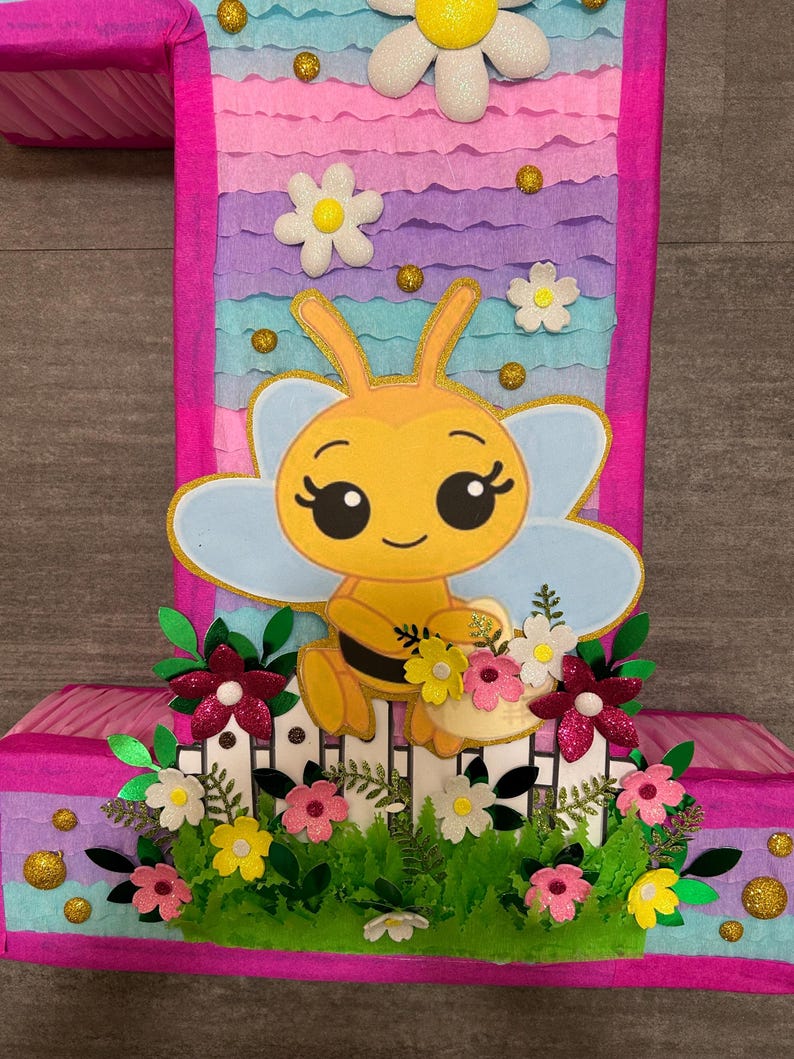 Pu&ograve; includere: Una pi&ntilde;ata rosa e blu a forma di numero uno con un'ape di cartone animato che tiene un cestino di fiori. L'ape &egrave; gialla con strisce nere e ha occhi grandi. La pi&ntilde;ata &egrave; decorata con fiori e glitter.