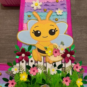 Pu&ograve; includere: Una pi&ntilde;ata rosa e blu a forma di numero uno con un'ape gialla che tiene un cestino di fiori. L'ape ha occhi grandi e un sorriso. La pi&ntilde;ata &egrave; decorata con fiori e foglie verdi.