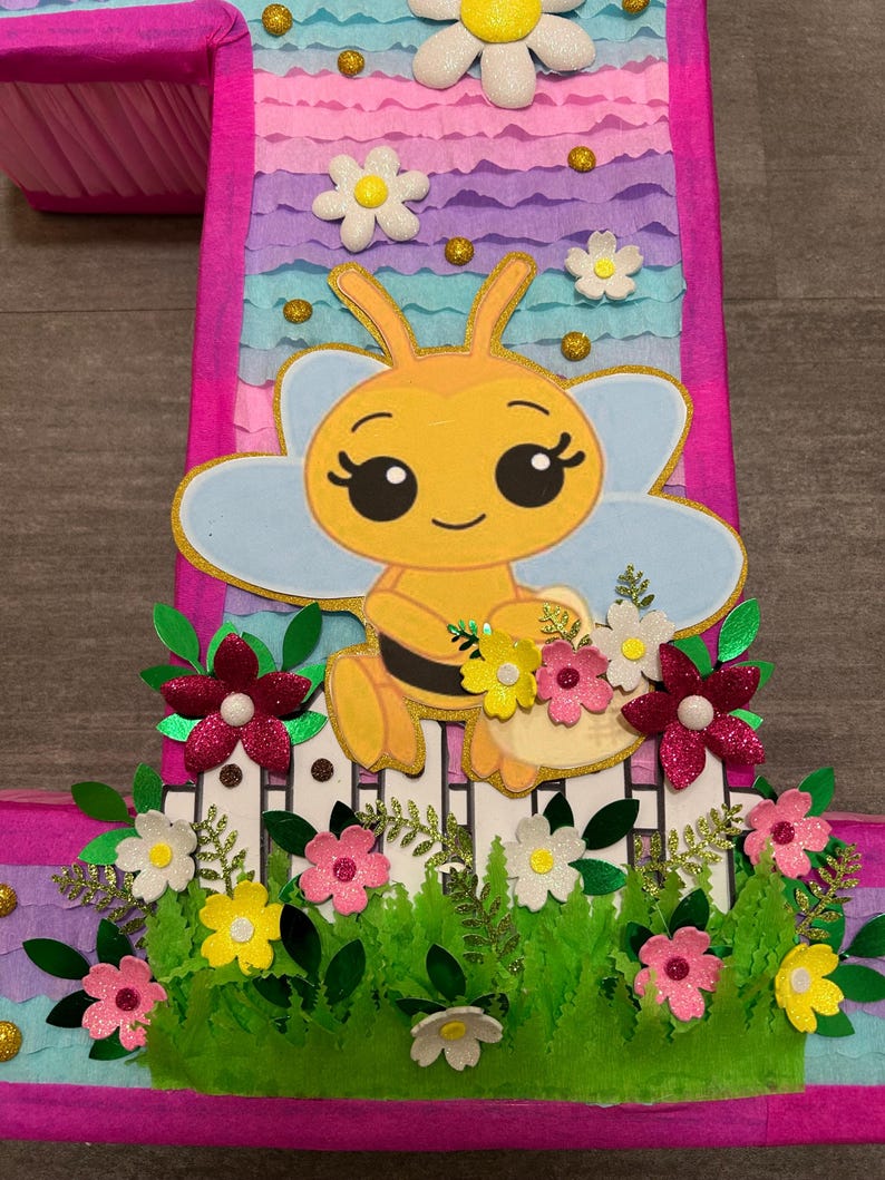 Pu&ograve; includere: Una pi&ntilde;ata rosa e blu a forma di numero uno con un'ape gialla che tiene un cestino di fiori. L'ape ha occhi grandi e un sorriso. La pi&ntilde;ata &egrave; decorata con fiori e foglie verdi.