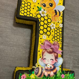 May include: A yellow and black number one pi&ntilde;ata with a bee and a girl with a pink bow. The pi&ntilde;ata is decorated with honeycomb and flowers.
