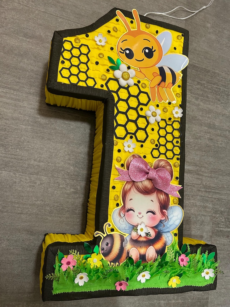 May include: A yellow and black number one pi&ntilde;ata with a bee and a girl with a pink bow. The pi&ntilde;ata is decorated with honeycomb and flowers.