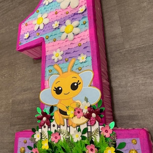 Pu&ograve; includere: Una pi&ntilde;ata a forma di numero uno rosa e blu con un design a forma di ape. La pi&ntilde;ata &egrave; decorata con fiori, glitter e una staccionata bianca.