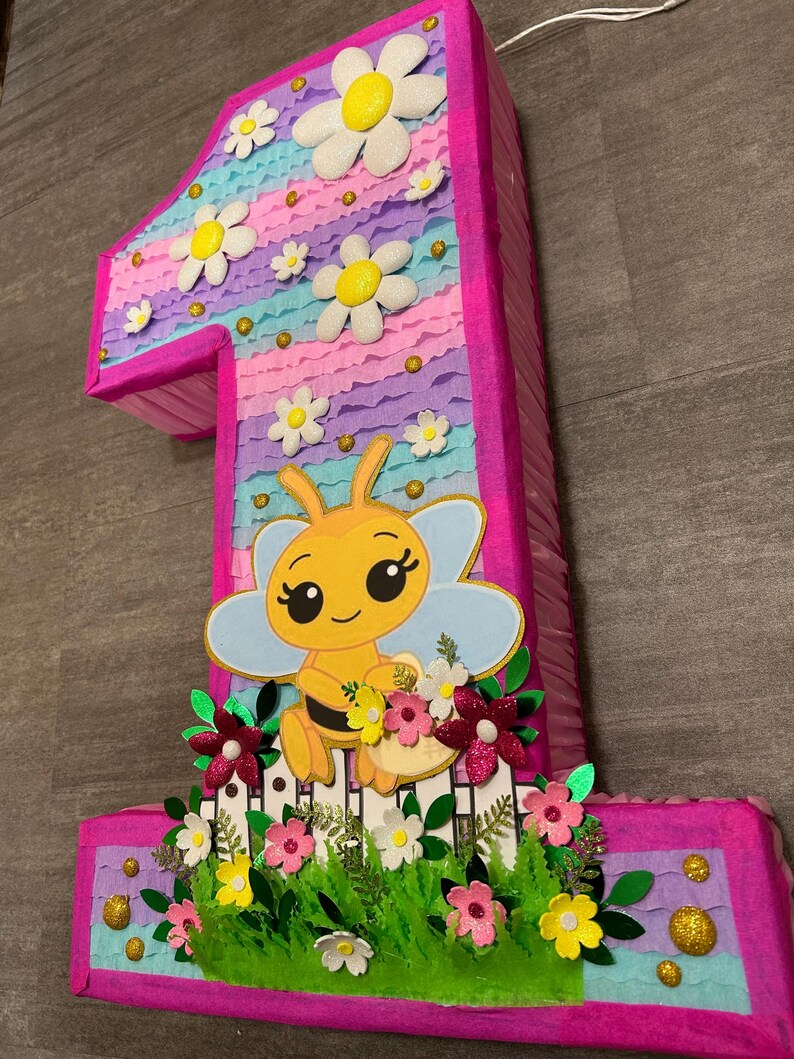 Pu&ograve; includere: Una pi&ntilde;ata a forma di numero uno rosa e blu con un design a forma di ape. La pi&ntilde;ata &egrave; decorata con fiori, glitter e una staccionata bianca.