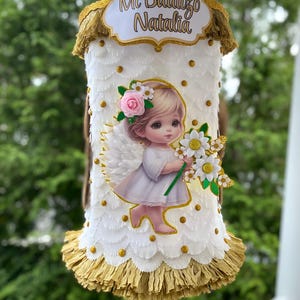 May include: White and gold pinata with a white angel holding a bouquet of white flowers. The pinata has a sign that says "Mi Bautizo Natalia".