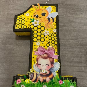 May include: A yellow number one pinata with a bee and a girl dressed as a bee. The pinata is decorated with honeycomb and flowers.