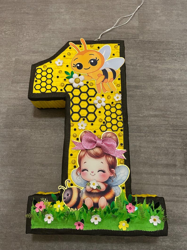 May include: A yellow number one pinata with a bee and a girl dressed as a bee. The pinata is decorated with honeycomb and flowers.