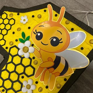 May include: A yellow and black bee pinata with a honeycomb pattern and white flowers. The bee has large eyes and a smiling face.