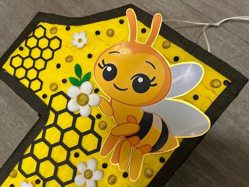 May include: A yellow and black bee pinata with a honeycomb pattern and white flowers. The bee has large eyes and a smiling face.