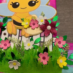 Pu&ograve; includere: Una pi&ntilde;ata rosa e blu a forma di numero uno, decorata con un'ape dei cartoni animati, fiori ed erba verde.