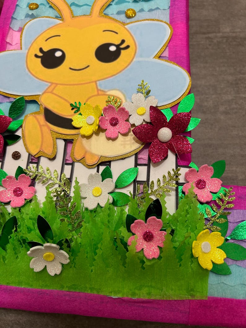 Pu&ograve; includere: Una pi&ntilde;ata rosa e blu a forma di numero uno, decorata con un'ape dei cartoni animati, fiori ed erba verde.