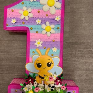 Pu&ograve; includere: Una pi&ntilde;ata a forma di numero uno rosa e blu con un'ape e dei fiori. La pi&ntilde;ata &egrave; decorata con margherite bianche e glitter dorato.