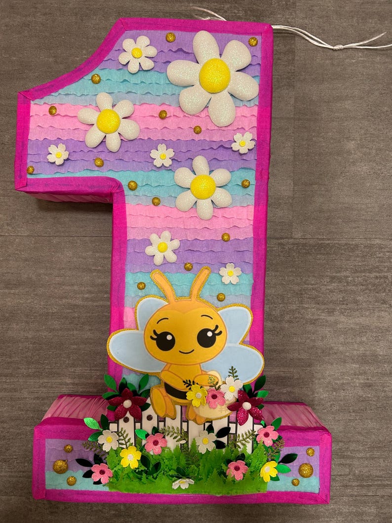 Pu&ograve; includere: Una pi&ntilde;ata a forma di numero uno rosa e blu con un'ape e dei fiori. La pi&ntilde;ata &egrave; decorata con margherite bianche e glitter dorato.