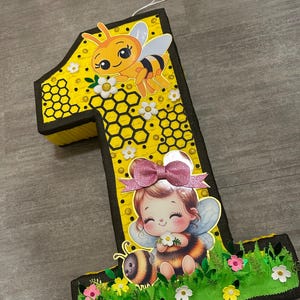May include: A yellow and black number one shaped pinata with a bee and a girl with bee wings. The pinata is decorated with honeycomb and flowers.