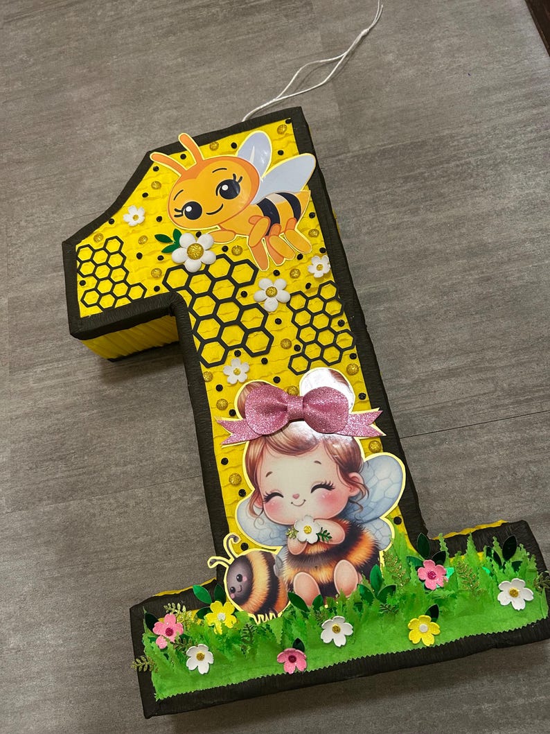 May include: A yellow and black number one shaped pinata with a bee and a girl with bee wings. The pinata is decorated with honeycomb and flowers.