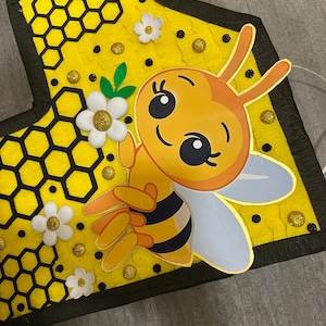 May include: A yellow felt bee with black stripes and white wings, holding a white flower with a yellow center. The bee is on a yellow background with a black honeycomb pattern and black dots.