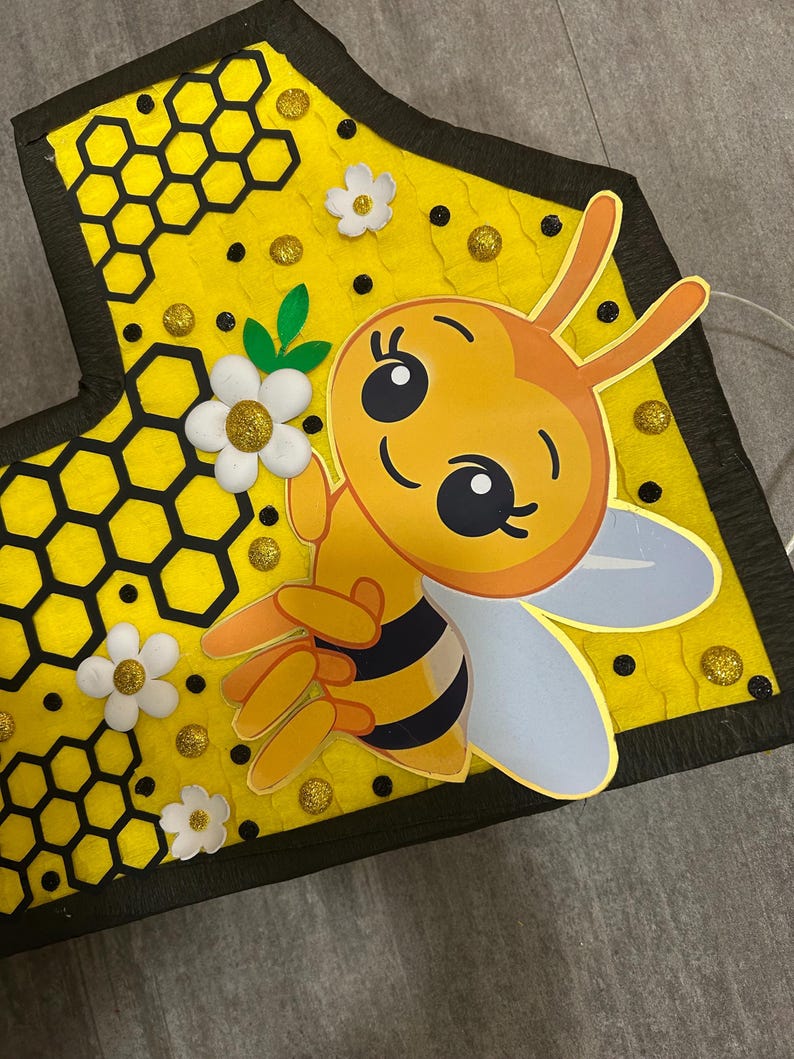 May include: A yellow felt bee with black stripes and white wings, holding a white flower with a yellow center. The bee is on a yellow background with a black honeycomb pattern and black dots.