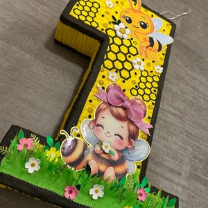 May include: A yellow and black number one shaped pinata with a bee and a girl with wings and a flower. The pinata is decorated with honeycomb and flowers.