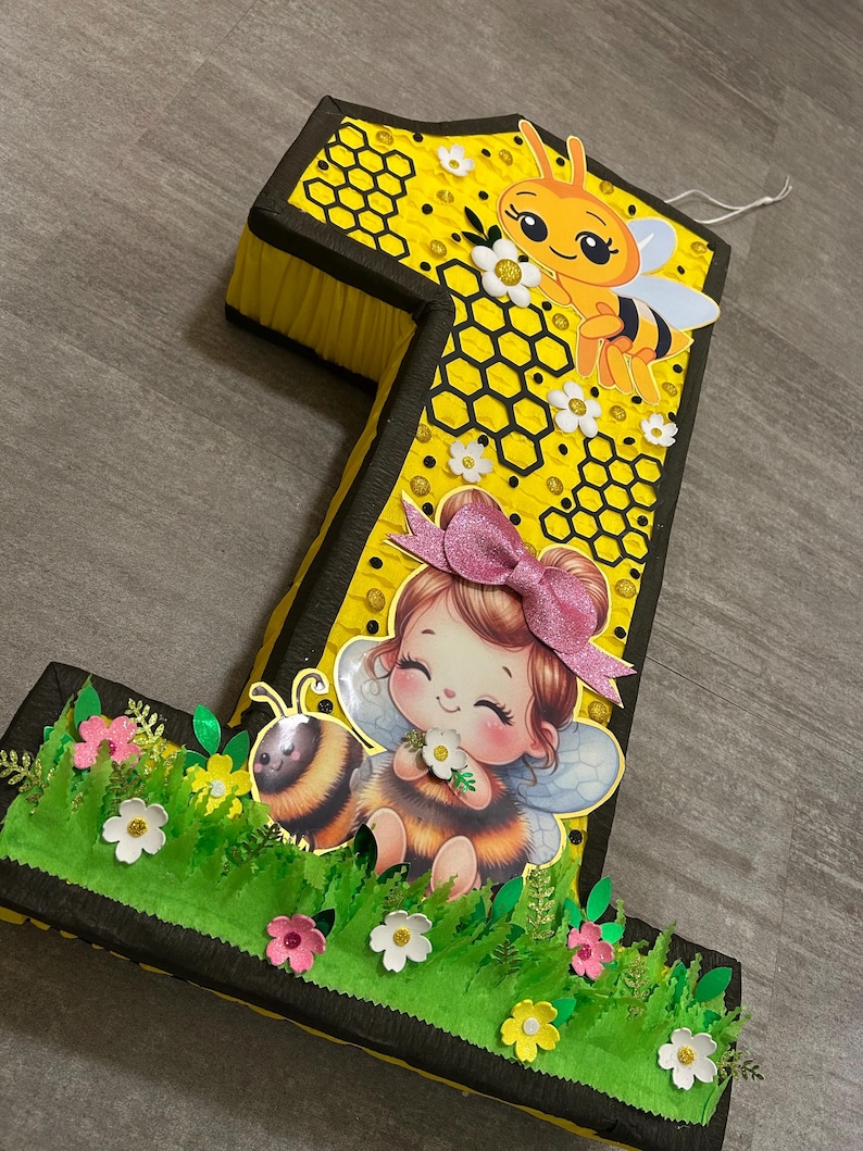 May include: A yellow and black number one shaped pinata with a bee and a girl with wings and a flower. The pinata is decorated with honeycomb and flowers.