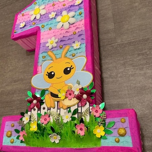 Pu&ograve; includere: Una pi&ntilde;ata a forma di numero uno rosa e blu con un'ape e dei fiori. La pi&ntilde;ata &egrave; decorata con margherite bianche, carta velina rosa e blu ed erba verde.