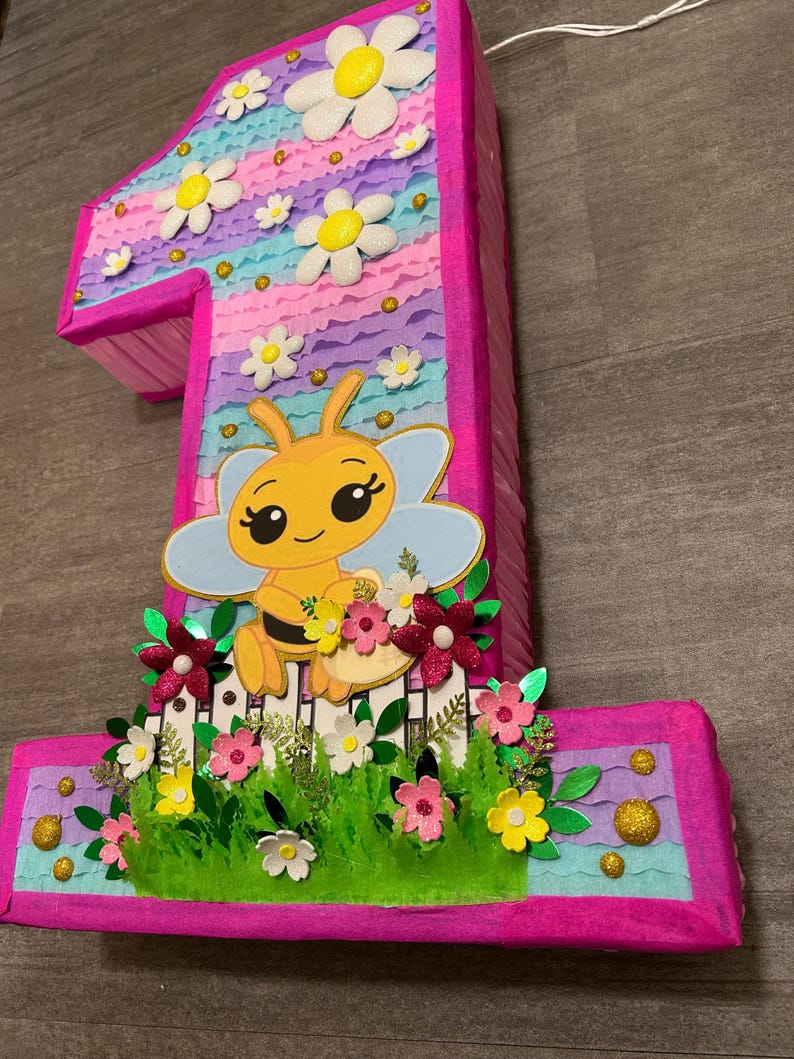 Pu&ograve; includere: Una pi&ntilde;ata a forma di numero uno rosa e blu con un'ape e dei fiori. La pi&ntilde;ata &egrave; decorata con margherite bianche, carta velina rosa e blu ed erba verde.