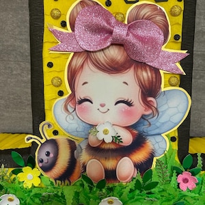 May include: A yellow and black honeycomb-patterned cardboard cutout of the number one with a cartoon bee girl wearing a pink bow and wings. The bee girl is holding a white flower and a bee is sitting next to her. The cutout is decorated with green leaves, yellow flowers, and pink flowers.