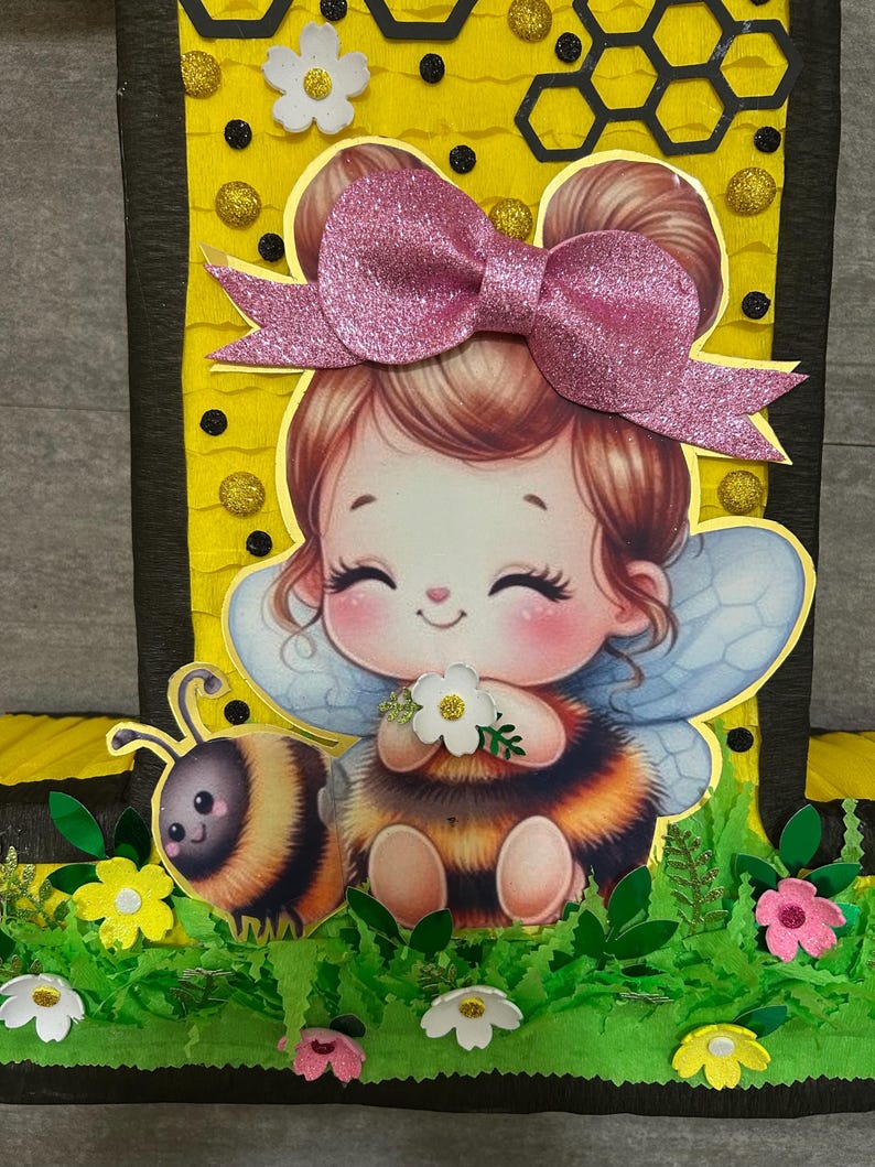 May include: A yellow and black honeycomb-patterned cardboard cutout of the number one with a cartoon bee girl wearing a pink bow and wings. The bee girl is holding a white flower and a bee is sitting next to her. The cutout is decorated with green leaves, yellow flowers, and pink flowers.
