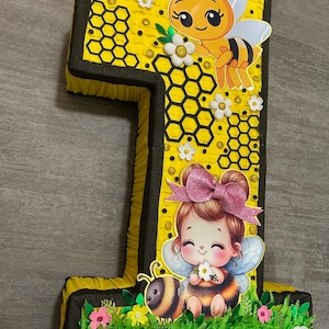 May include: A yellow and black number one pi&ntilde;ata with a bee and a girl with wings and a flower. The pi&ntilde;ata is decorated with honeycomb, flowers, and glitter.