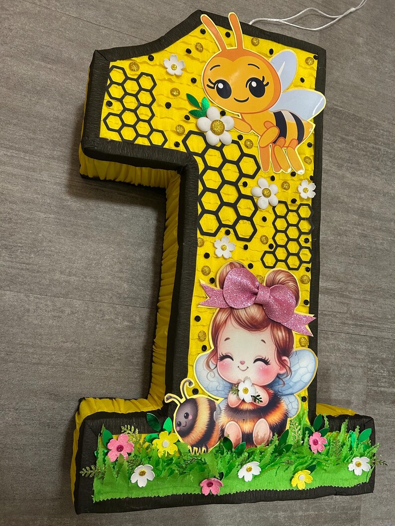 May include: A yellow and black number one pi&ntilde;ata with a bee and a girl with wings and a flower. The pi&ntilde;ata is decorated with honeycomb, flowers, and glitter.